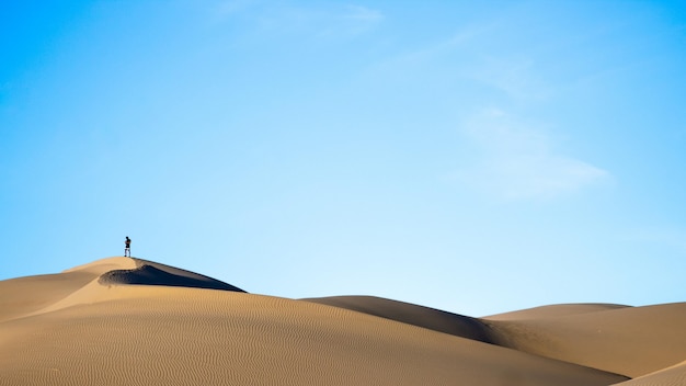 Horizontal shot of a person standing on sand dunes in a desert with the blue sky in the back