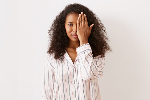 Horizontal shot of playful adorable young African American female with voluminous black hair and brown tanned skin posing at blank wall in striped satin pajamas, covering one eye with hand