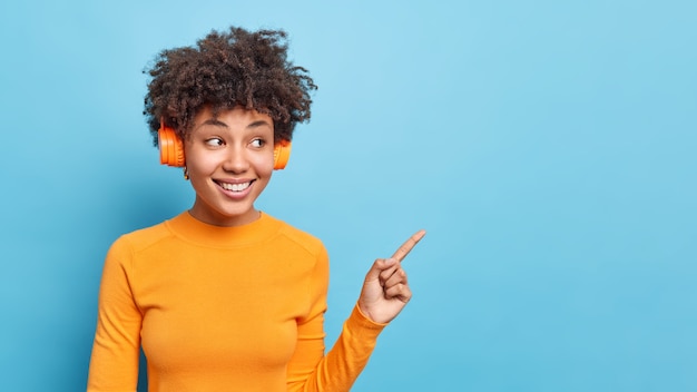 Free photo horizontal shot of pleasant looking afro american woman listens audio track dressed in casual orange jumper points away on blue copy space suggests to check out promo demonstrates announcement