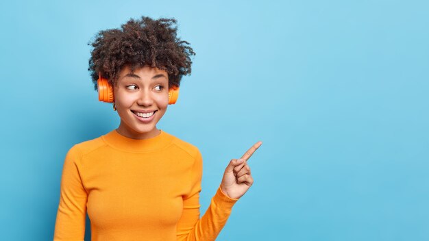 즐거운 찾고 아프리카 계 미국인 여자의 가로 샷 파란색 복사 공간에 캐주얼 오렌지 점퍼 포인트를 입고 오디오 트랙을 듣고 프로모션을 확인하는 것이 좋습니다 발표 발표