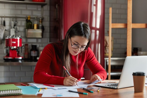 Горизонтальный снимок симпатичной деловой женщины, которая работает с бумагами дома, готовит отчет, заполняет информацию, сидит перед открытым ноутбуком на кухне.