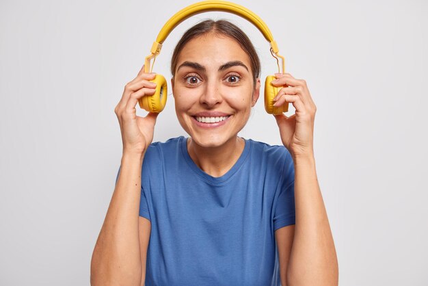 Horizontal shot of pleasant looking cheerful woman takes off headphones listens music with loud sound smiles toothily dressed casually isolated over white background tries to hear what you said