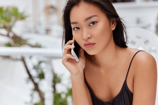 Free photo horizontal shot of pleasant looking female with attractive appearance, talks via smart phone with serious expression, sits at outdoor cafeteria, waits for business partner. people, technology, rest