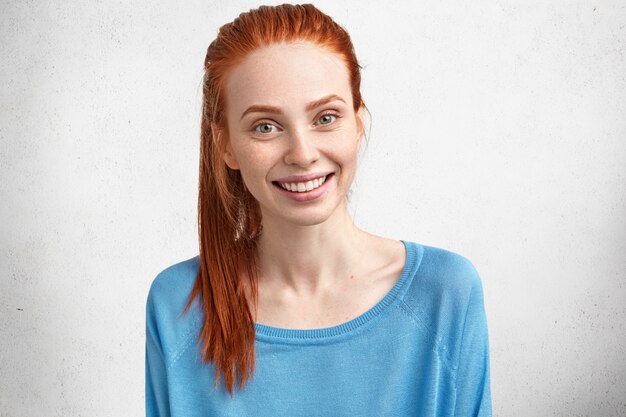 Horizontal shot of pleasant looking glad female with freckled skin and positive smile, dressed in blue sweater, isolated over concrete wall.