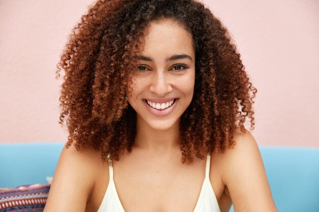 Horizontal shot of pleased attractive young female model with curly dark hair, pleasant smile on face
