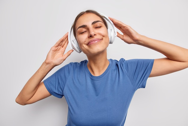 Horizontal shot of pleased dreamy young woman wears stereo headphones on ears enjoys listening music keeps eyes closed wears casual blue t shirt isolated over white background People and hobby