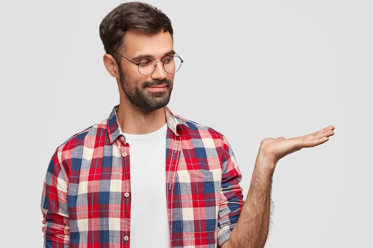 Free Photo horizontal shot of pleased man has thick bristle, raises palm, pretends holding something, wears checkered shirt with spectacles, stands against white wall