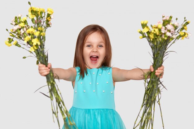 満足している小さな子供の水平方向のショットは、2つの花束を保持し、口を開け、幸福と叫び、白い壁に分離された青いドレスを着ています。