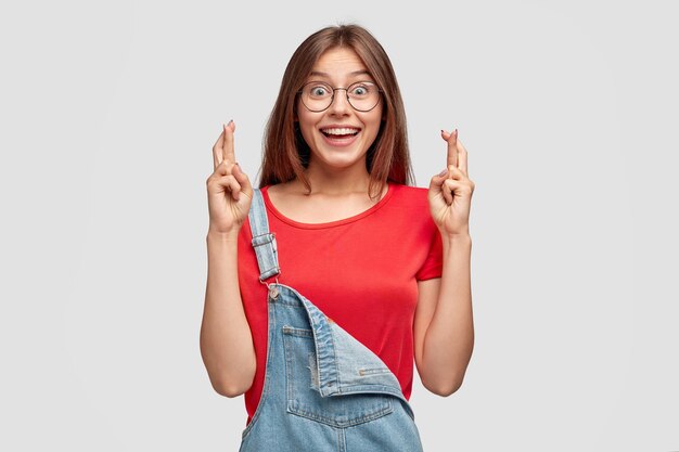 Horizontal shot of pleased smiling teenager keeps fingers crossed, believes in good fortune before passing exam