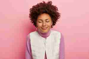 Free photo horizontal shot of pleased woman has curly hairstyle, eyes closed, calm expression, wears round earrings and casual wear, poses against pink background.