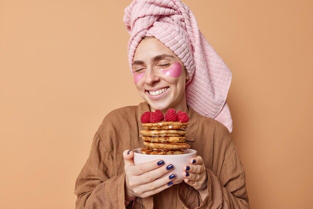Horizontal shot of pleased young woman closes eyes from pleasure holds yummy pancakes with marple syrup smiles pleasantly dressed in slumber suit undergoes beauty procedures after taking shower