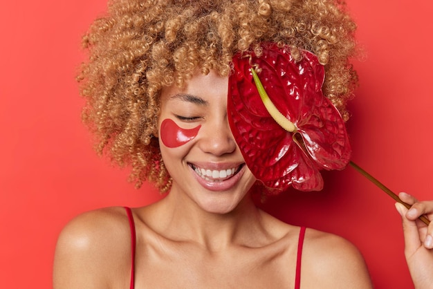 Horizontal shot of pleased young woman smiles happiy undergoes beauty procedures covers eye with red calla applies collagen patches poses indoor dressed casually. skin care and wellness concept
