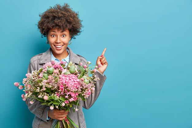 Horizontal shot of positive curly haired woman dressed in grey jacket points away on blank space holds bunch of flowers celebrates birthday isolated over blue wall