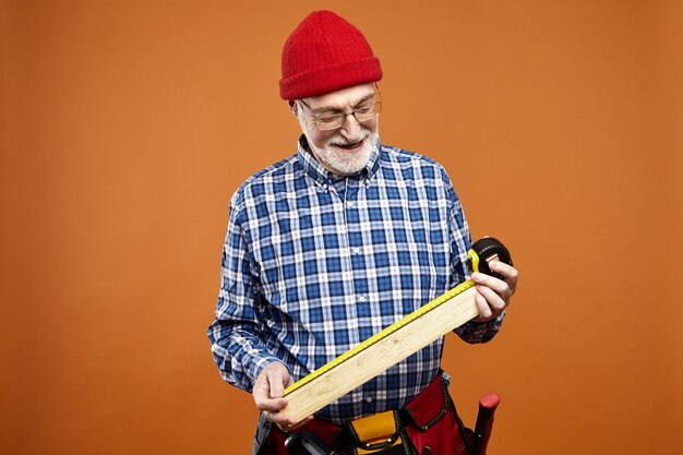 Horizontal shot of positive skilled elderly European craftsman wearing pveralls and eyeglasses holding piece of future wooden furniture, measuring its size and smiling, enjoying manual work