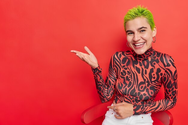 Horizontal shot of positive trendy girl involved in teen subculture grabs your attention to blank space keeps palm raises shows advertising content wears turtleneck white jeans isolated over red wall