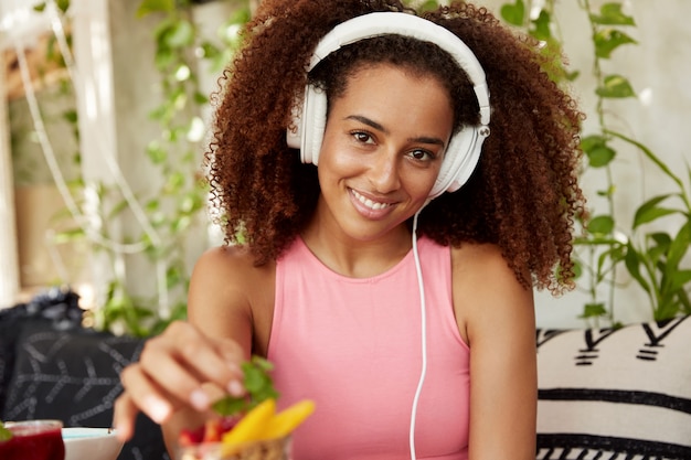 Inquadratura orizzontale della donna positiva ascolta nuova musica in cuffie moderne mentre aspetta un amico nella caffetteria, mangia dessert alla frutta, gode di un buon riposo. gli studenti afroamericani ricreano dopo le lezioni