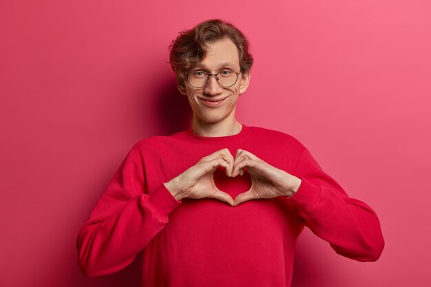 Horizontal shot of positve passionate handsome man speaks about own feelings, makes heart gesture over chest, expresses sympathy and love, smiles positively, wears round spectacles and jumper