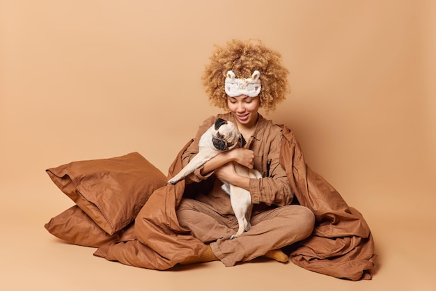 Free photo horizontal shot of pretty cheerful woman plays with favorite pug dog stay in bed wears pajama sits in lotus pose wrapped in blanket isolated over beige background rest domestic animals people