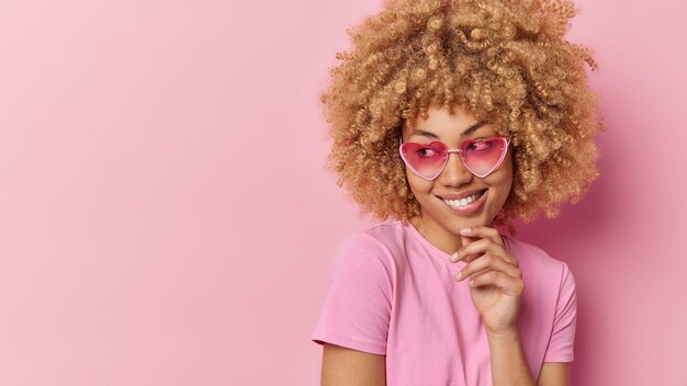 Horizontal shot of pretty curly haired woman smiles toothily keeps hand on chin wears heart sunglasses and casual t shirt isolated over pink background blank empty space for your advertisement