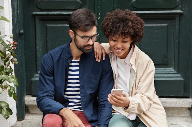 Free photo horizontal shot of pretty dark skinned girl shows her photos on cell phone to best friend