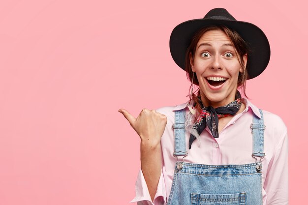 Horizontal shot of pretty female gardener with positive expression, points aside with thumb, dressed in casual overalls and black hat