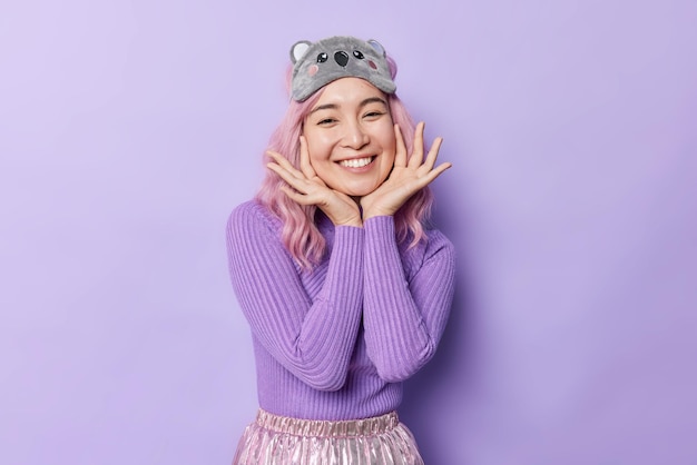 Free photo horizontal shot of pretty happy woman touches face gently smiles broadly has glad expression wears casual turtleneck and sleepmask poses against purple background positive emotions concept