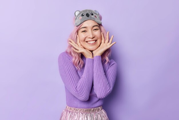 Horizontal shot of pretty happy woman touches face gently smiles broadly has glad expression wears casual turtleneck and sleepmask poses against purple background Positive emotions concept