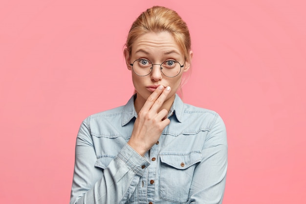 Horizontal shot of pretty light haired female looks with bewilderment directly at camera, wears round spectacles keeps fingers on lips, has focused look at interlocutor listens attentively information