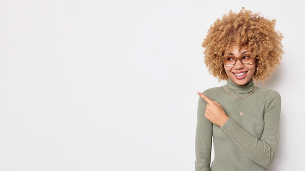 Horizontal shot of pretty positive woman with curly hair points away at empty space shows place for your advertisement suggests to click on link dressed casually isolated over white background