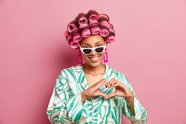 かなり満足しているアフロアメリカ人女性の水平ショットは、ヘアスタイルを作るためにヘアローラーを適用しますシルクのガウンサングラスとイヤリングはピンクの壁に隔離されたハートサインを形作ります