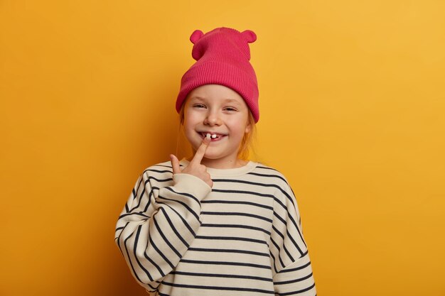 Горизонтальный снимок довольно маленькой девочки указывает на ее новый зуб, заботится о зубах, носит стильную одежду, забавное выражение лица, хихикает в помещении, изолирована на ярко-желтой стене