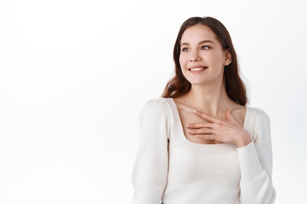 Horizontal shot of pretty smiling female with charming smile, touches heart, being moved to be praised by someone, wears casual clothes, stands indoor on white wall