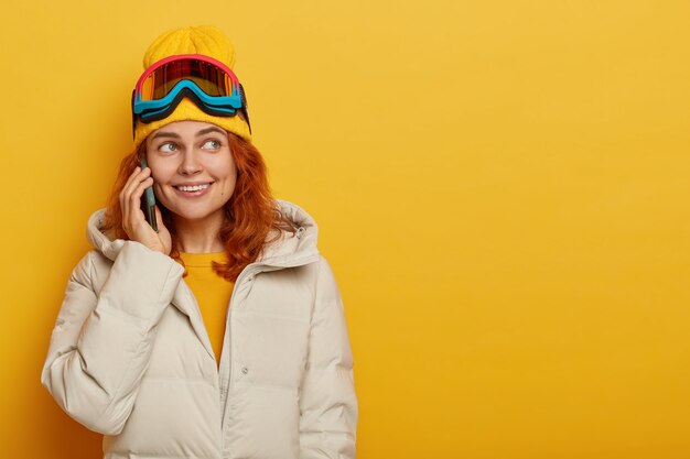 Horizontal shot of pretty smiling girl skier calls relatives vis smartphone, tells about her winter vacation, wears snowboarding goggles