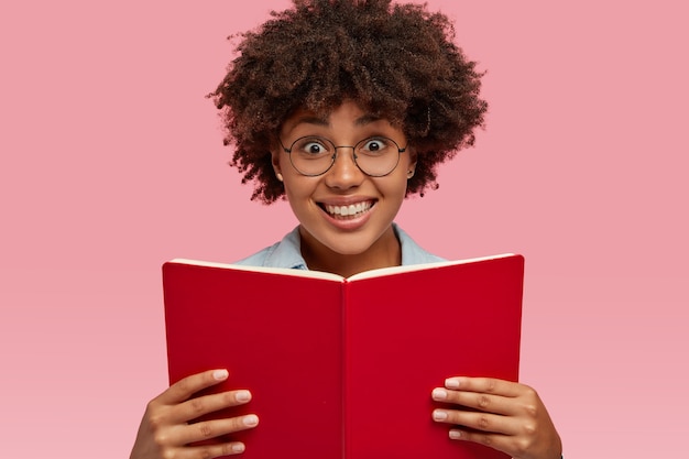 Horizontal shot of pretty smiling girl with pleasant facial expression, carries textbook, wears spectacles, prepares for examination, isolated over pink wall. People, ethnicity and literacy concept