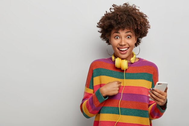 Horizontal shot of pretty woman points at herself, happy being chosen for promotion, holds modern cellphone, yellow headphones on neck