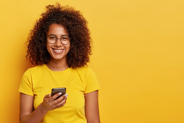 얼굴에 유쾌한 미소를 지닌 예쁜 여자의 가로 샷, 휴대 전화에서 온라인 커뮤니케이션을 즐기고, 알림을 읽고, 둥근 안경과 노란색 티셔츠를 입습니다. 기술과 사람 개념