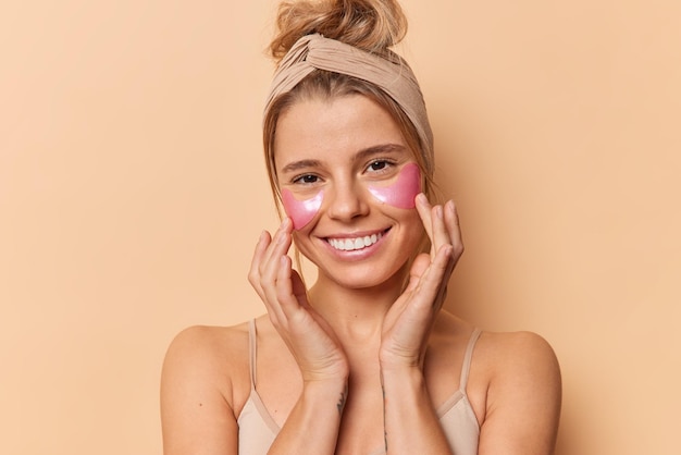 Horizontal shot of pretty young European woman touchs face gently enjoys softness of skin applies pink hydrogel patches under eyes wears headband isolated over beige background smiles happily