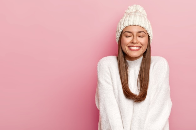 Horizontal shot of pretty young female with dark hair, keeps eyes shut, smiles pleasantly, shows white perfect teeth, enjoys comfort in new bought sweater, warm hat