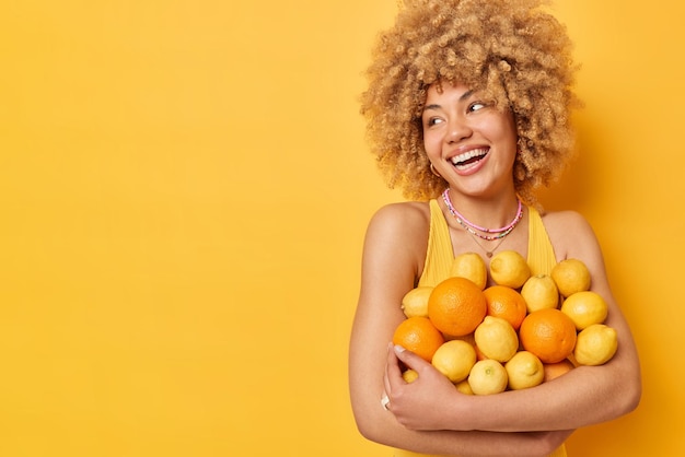 예쁜 젊은 여성의 수평 사진은 신선한 즙이 많은 오렌지와 과일을 껴안고 노란색 배경 복사 공간에 격리된 얼굴에 기쁜 표정을 짓고 있습니다.