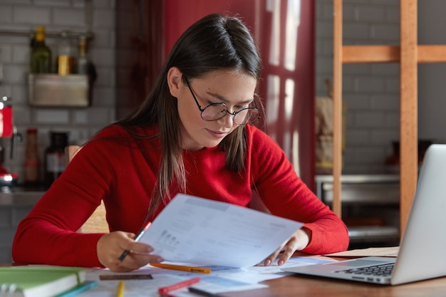 Horizontal shot of project manager in spectacles and red juumper, looks attentively at documents, thinks how attract clients and increase income, poses against kitchen interior with portable laptop