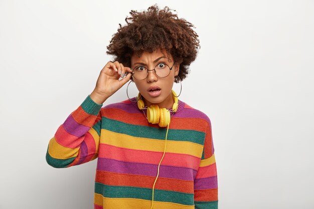 Horizontal shot of puzzled curly Afro American woman touches frame of spectacles, looks surprisingly, hears something amazing, wears striped mutlicolored jumper