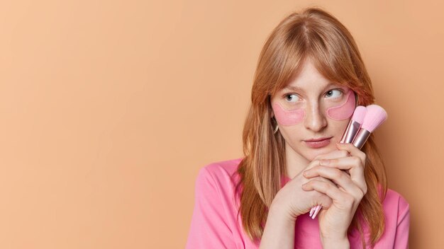 Horizontal shot of redhead girl with freckled skin applies hydrogel pink patches under eyes holds cosmetic brushes focused away isolated over beige background empty space for your promotion