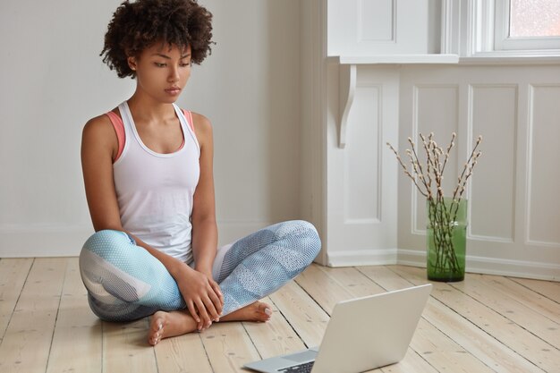 Horizontal shot of relaxed dark skinned woman sits in lotus pose, watches training video, learns yoga, dressed in casual clothes, poses on wooden floor in empty room. Technology and rest concept