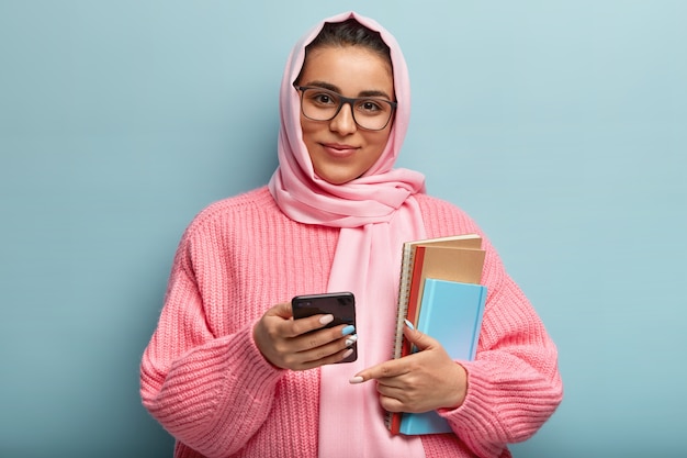 Horizontal shot of satisfied college student uses new cool app on cell phone, carries notepad for writing notes, wears spectacles, silk scarf and knitted sweater, isolated over blue wall.