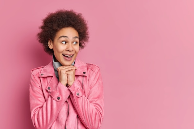 Free photo horizontal shot of satisfied curly haired woman keeps hands under chin smiles gladfully looks with interest aside