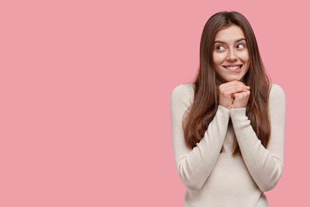 Horizontal shot of satisfied European woman holds hands under chin, bites lower lip, looks with mysterious expression aside, satisfied to recieve something good