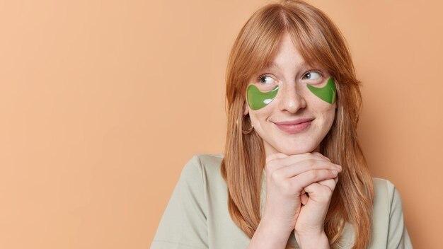 Horizontal shot of satisfied freckled girl with ginger hair keeps hands under chin looks happily away applies green hydrogel patches to remove puffiness isolated over beige background copy space