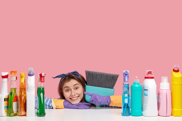 Horizontal shot of satisfied housemaid wears headband, points at detergents, carries broom closely, likes its perfect effect