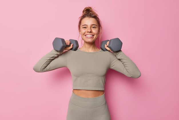 Horizontal shot of satisfied motivated sportswoman has regular training raises arms with dumbbells does exercises for muscles dressed in sportswear smiles toothily isolated on pink wall. Sport concept