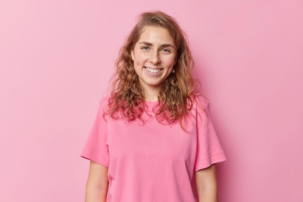 Горизонтальный снимок довольной улыбающейся женщины с темными волнистыми волосами, которая в хорошем настроении проводит свободное время дома в повседневной футболке, изолированной на розовом фоне. Концепция положительных человеческих эмоций